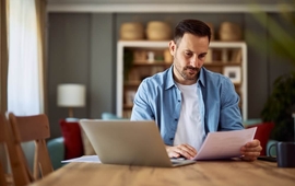 A concentrated adult man reading and analyzing a document while sitting in front of a laptop (1).jpg