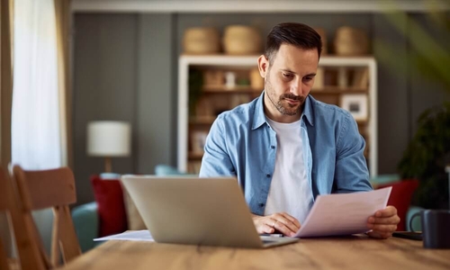 A concentrated adult man reading and analyzing a document while sitting in front of a laptop (1).jpg