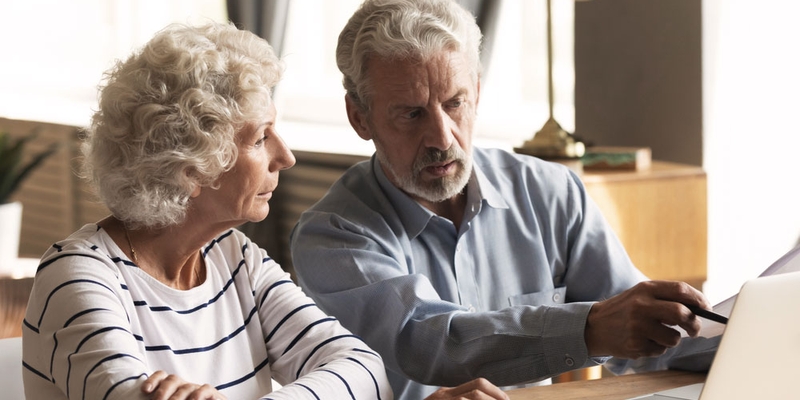 A senior woman sits next to a senior man and points at a computer with a worried look on his face