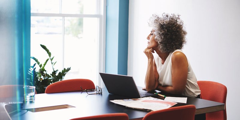 An older professional woman gazes out the window while taking a break from working on her laptop at home