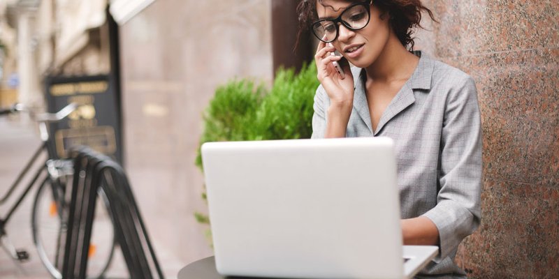 A professional woman in trendy glasses sits outside at a table while working on her laptop and talking on the phone