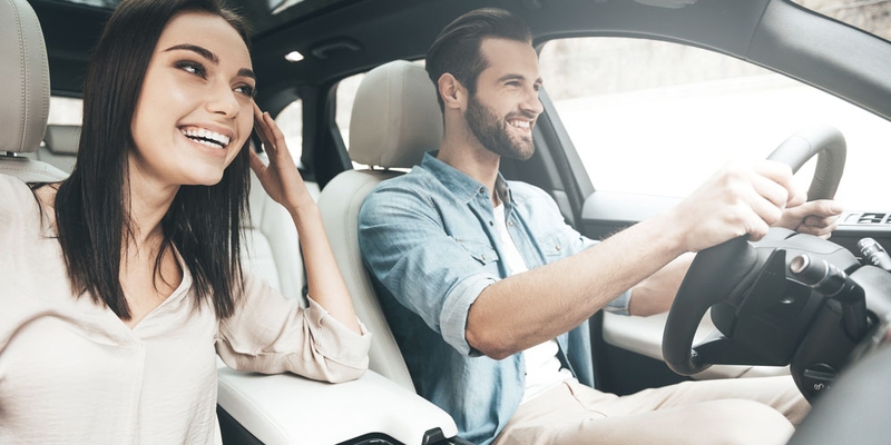 A male driver sits smiling with a female passenger on a bright day