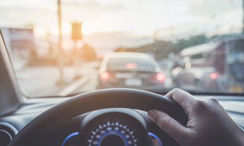 A hand is seen gripping the steering wheel while the car sits in traffic